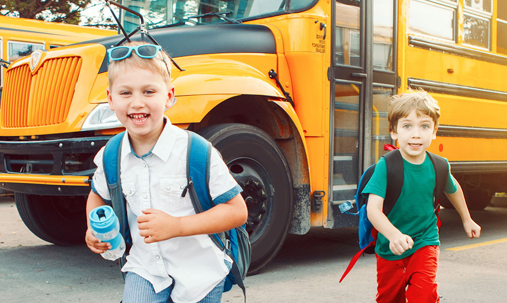 Image of a child wearing a backpack smiling with a smaller child behind him and a school bus in the background